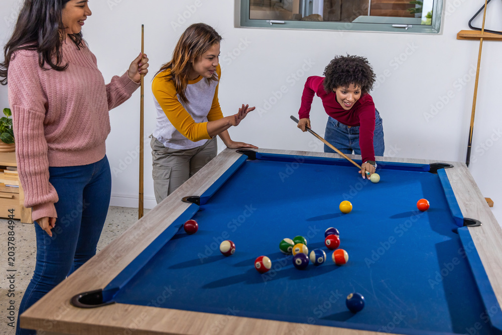 Fototapeta premium Diverse female friends lining up pool shot on blue-felt table in game room holding cues