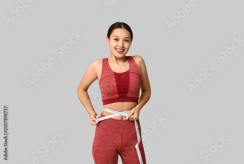 Young woman in sportswear measuring her waist on light background. Weight los...