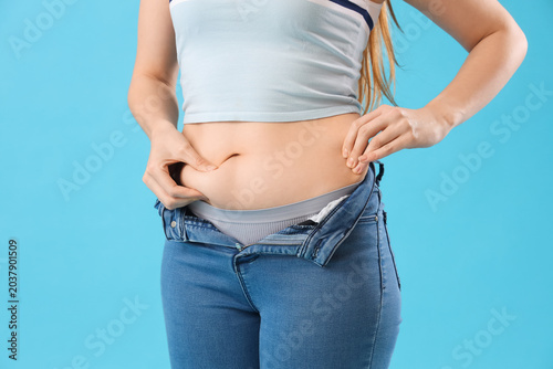 Young woman in tight jeans on blue background, closeup. Weight gain concept