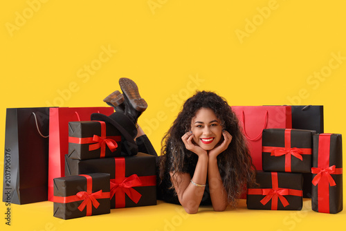Beautiful African-American woman with shopping bags and gift boxes on yellow ...