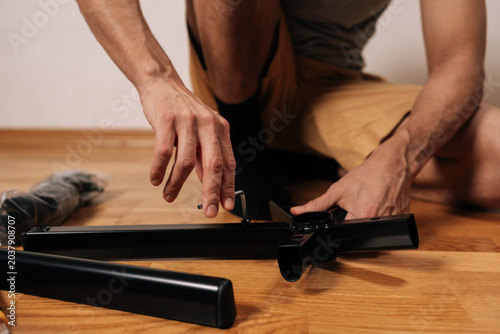 Master male assembling new office chair furniture, carefully using allen key to attach legs to central base while kneeling on wooden floor during home project.