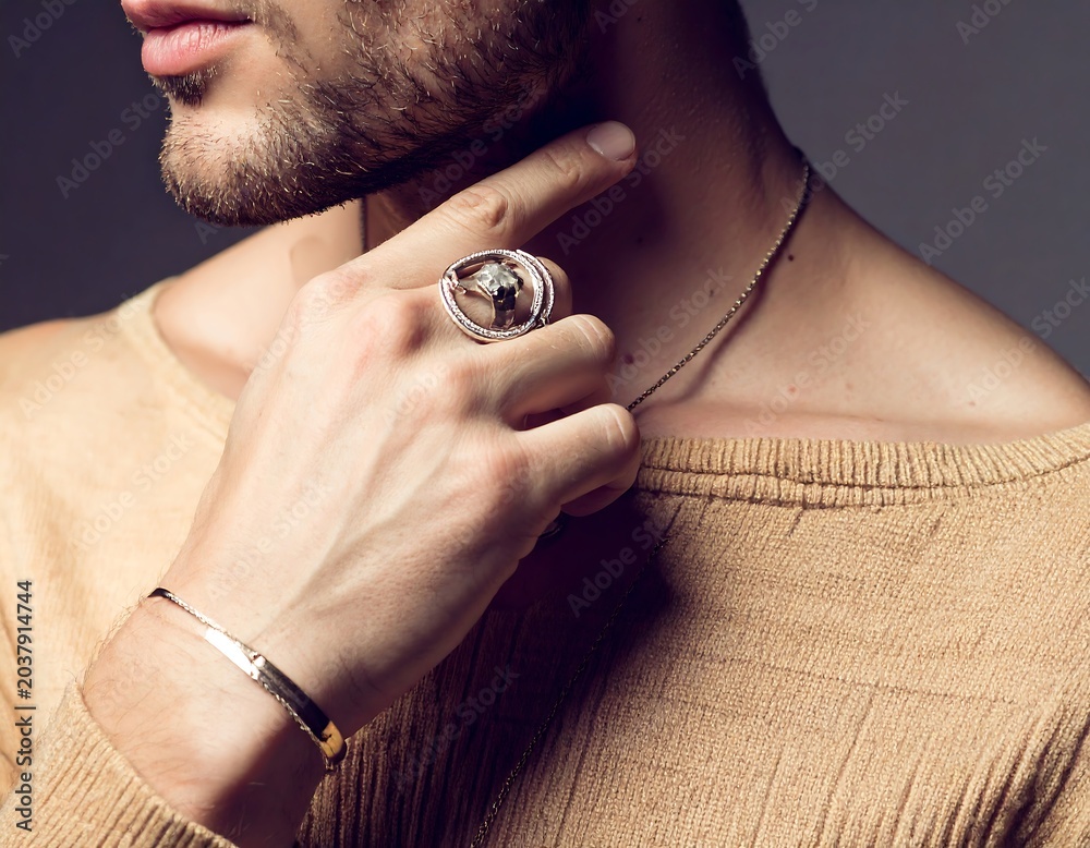Fototapeta premium Close-up of a man in tan clothing, adorned with a ring and bracelet, touching his neck, with a necklace