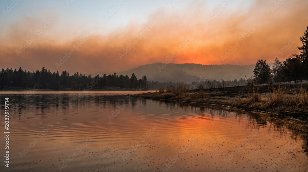 Fototapeta premium Smoky orange sunset over lake with reflection and misty mountains 