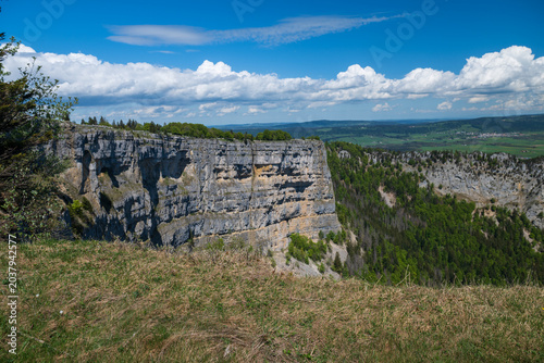 Panoramic Swiss Jura landscape with limestone amphitheater, wildflower meadow, forests, and dramatic clouds under blue sky at Creux du Van.