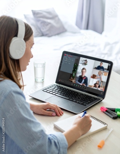 A young woman wearing headphones participates in a group video conference on her laptop at home. Perfect for concepts of online education, remote work, and modern elearning.