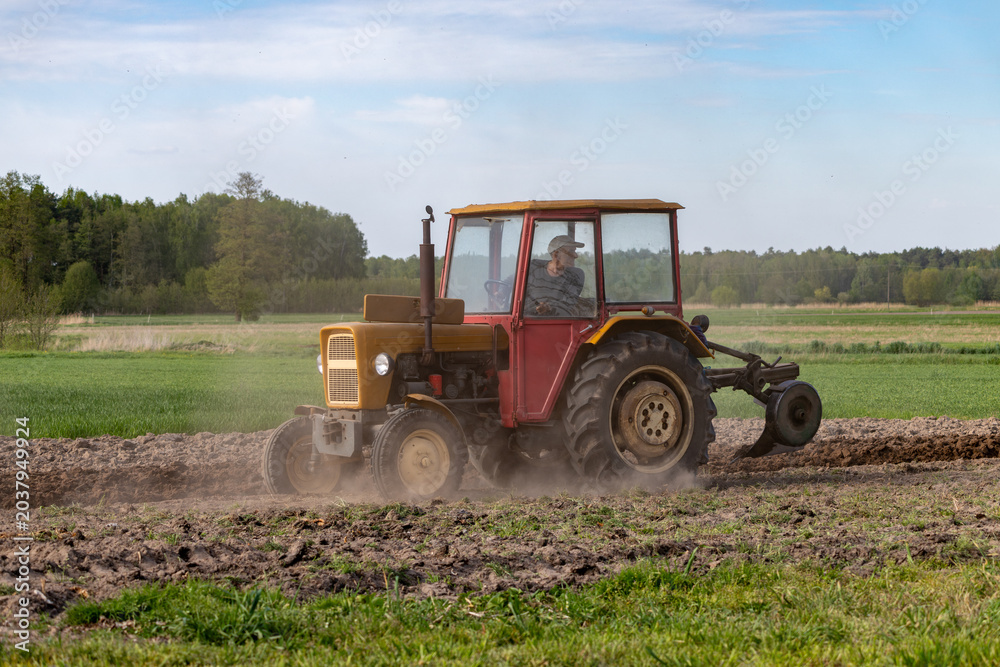 Fototapeta premium Old yellow tractor plowing agricultural field raising dust during spring seasonal farming work with green forest and blue sky in the background countryside