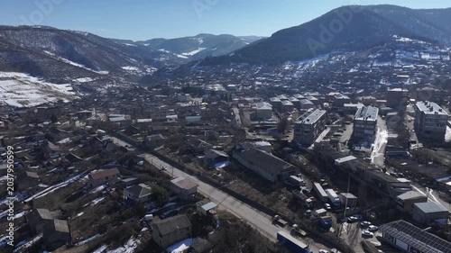 A stunning aerial drone shot flying over a scenic mountain village with houses and apartments nestled in a snowy valley under a clear blue sky in beautiful Armenia.

