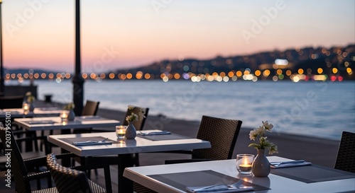 Empty waterfront restaurant tables set for dinner with city lights reflecting on the water at dusk