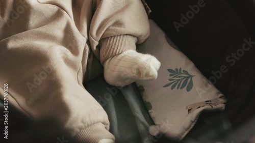 Static close up shot of a baby wearing a beige romper and white socks moving their legs while lying in a stroller with a patterned pillow.