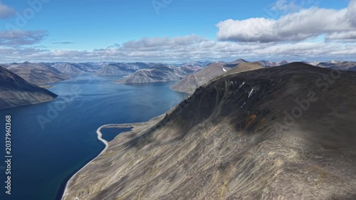 Aerial view past a steep cliff as the drone glides along the slope. Wide blue bay and mountain backdrop in daylight, Russia, Chukotka, Provideniya. Perfect for travel storytelling and tourism
