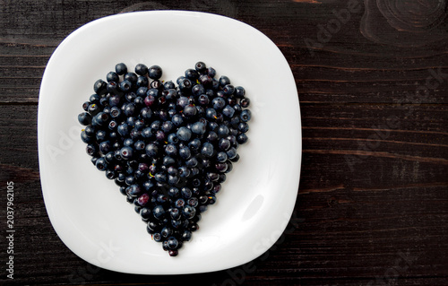 Fresh ripe blueberries arranged in a heart shape on a white plate over a dark wooden background.