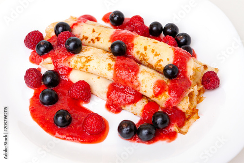 Sweet rolled crepes topped with red berry sauce and fresh raspberries with blackcurrants on a white plate.