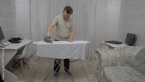 Man irons sheet. Gentleman meticulously presses bedsheet in comfortable living space during leisure. Man engages in household chore of ironing amidst cozy home setting and relaxing ambiance