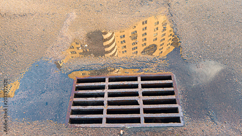 Fotografía Urban building reflecting in a water puddle on pavement, showing worn metal stor