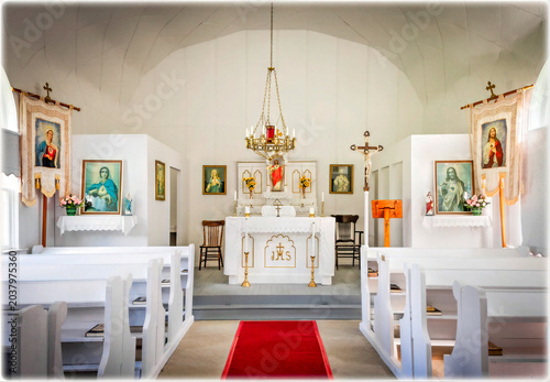 The image shows a quaint rural church interior with white pews and a red carpet leading to the altar. Various religious icons and images hang on the wall, completing the serene atmosphere.