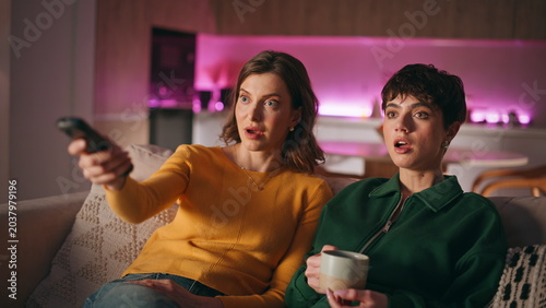 Sisters choosing tv movie switching channels sitting together cozy couch.