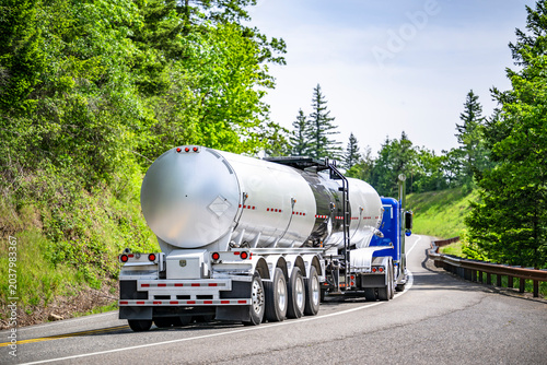 Bright blue big rig semi truck transporting liquid cargo in long tank semi trailer with four axels running on the winding road with hills and forest on the sides