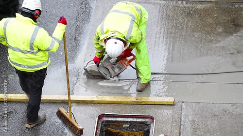 Two construction worker cutting concrete road slab with saw and broom nearby