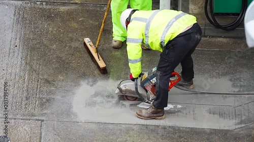 Worker cutting concrete slab with circular saw beside broom at road site