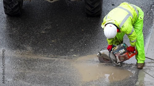 Worker in yellow vest cutting wet concrete road beside puddle with power saw