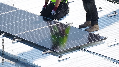 Worker placing solar panel on metal rooftop while colleague assists at site