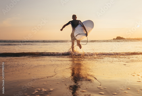 back view of active male surfer with surfboard running fast into ocean water on sandy beach during golden sunset in Sri Lanka representing water sports outdoor recreation travel adventure vacation