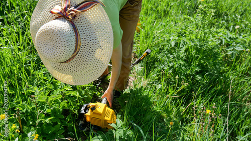 Obraz na płótnie Woman in sun hat using string trimmer to cut tall grass in outdoor rural field d