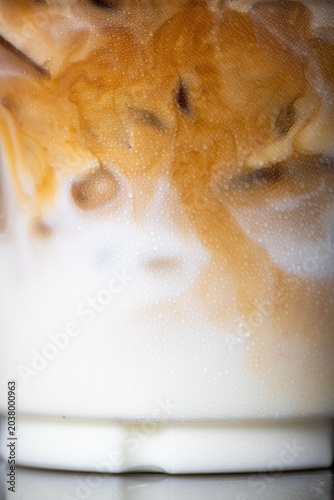 Close-up iced latte coffee showing beautiful layers of dark espresso swirling into white milk with ice cubes inside a plastic cup.