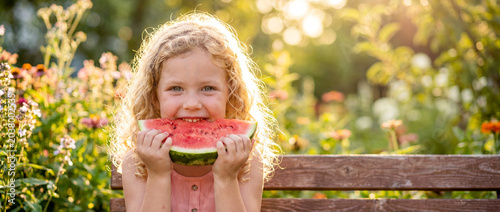 Curly blonde little girl is eating a fresh sweet watermelon slice outdoors on a beautiful summer day