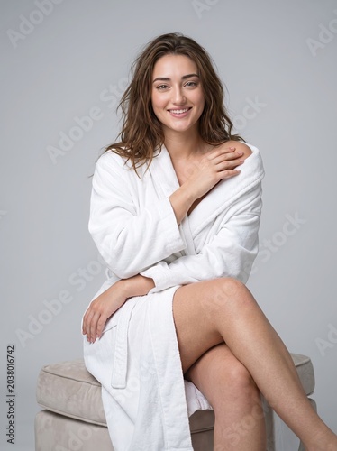 Radiant young woman with a warm smile, wearing a comfortable white bathrobe, exudes natural beauty and relaxation while posing gracefully in a clean studio setting, embodying self-care and wellness