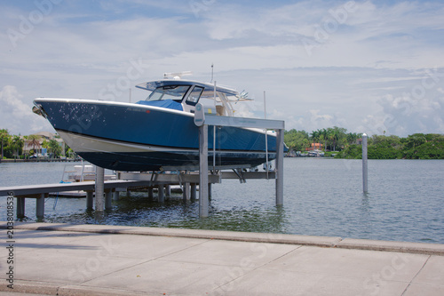 Obraz na plátne Luxury motorboat sits elevated on a boat lift beside a residential dock, overlooking calm waterfront with palm-lined shore, blue sky and summer clouds — a serene coastal leisure scene