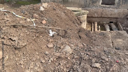 Exposed heating pipes outside panel apartment foundation repair site with large trench dirt and rubble around corroded metal pipes
