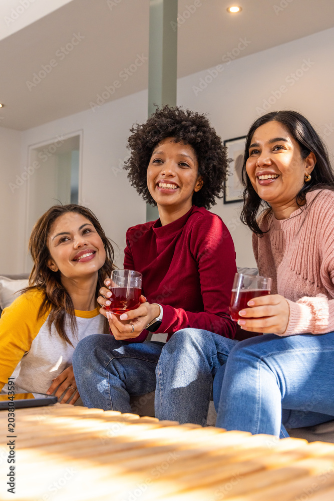 Naklejka premium Diverse female friends sitting on couch in living room holding red drinks near coffee table, tablet