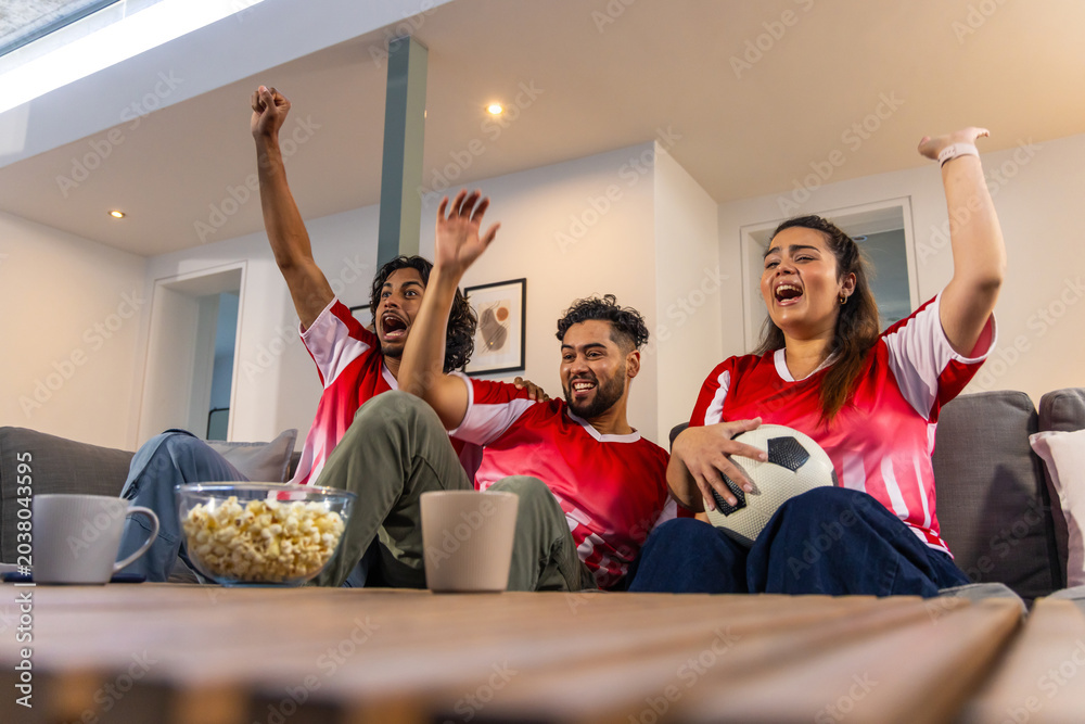 Naklejka premium Friends cheering on gray sofa in living room, wearing red jerseys with soccer ball and popcorn
