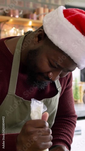 Vertical video: Squeezing piping bag, man icing holiday cookies in kitchen with Santa hat  and  apro