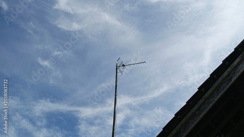 Stock image of a daytime blue sky filled with wispy white cirrus clouds, featuring a television antenna and house rooftop silhouette.
