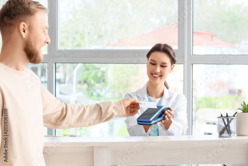 Young man with credit card paying via terminal at reception in clinic