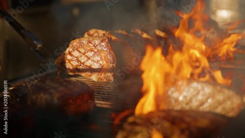 Premium steaks being prepared in super expensive restaurant as chef uses metal tongs to flip thick cuts on flaming grill. Slow motion footage captures dramatic fire and smoke in professional kitchen.