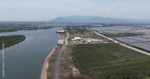 Backward aerial view of the tourist port in Kampot, featuring a modern, white building by the river, sprawling salt pans, and a distant mountain range.