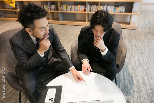 本棚のある社長室で資料を見ながら相談、ビジネスマンの打ち合わせ　契約書など行政書士や士業のイメージ困る考える悩む俯瞰のイメージ