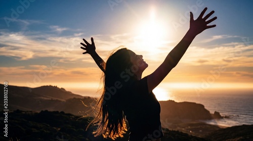 A young girl stands back-lit with arms open wide to a coastal golden sunset, her hair catching the warm light. The image evokes absolute freedom, joy, and hope.