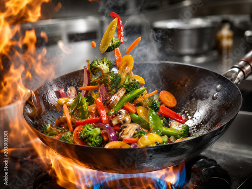 A high-energy shot of a vegetable stir-fry being prepared in a wok, featuring flames, steam, and professional cooking motion.