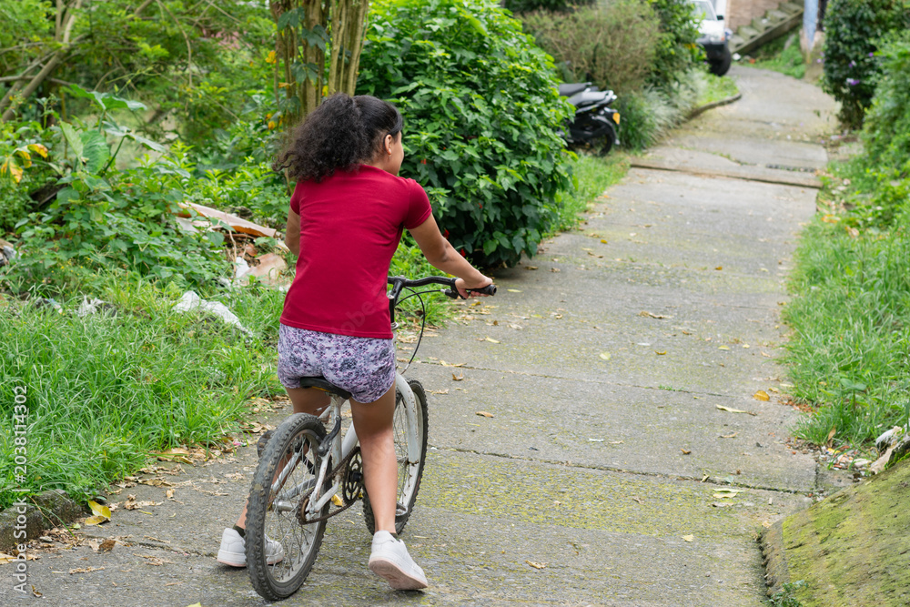 Fototapeta premium Young Latina girl riding bicycle downhill on asphalt street in a low income neighborhood outdoors