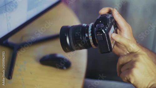 A person holds a DSLR camera with a lens, seemingly preparing to take a photo. In the background, a computer monitor and mouse sit on a wooden desk.