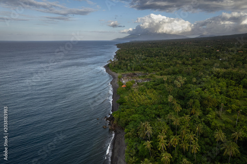 An aerial view follows the black sand coastline of Karangasem in eastern Bali, where ocean waves meet a volcanic shore lined with palm trees and tropical forest