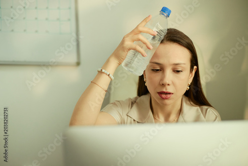 Fotografie Young adult Caucasian businesswoman cooling forehead with water bottle while working on laptop, showing heat stress and fatigue