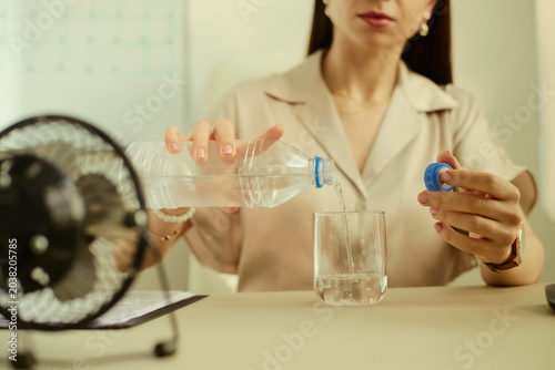 Obraz na płótnie Young adult woman pouring water into glass beside desk fan, demonstrating hydration during heat at workplace