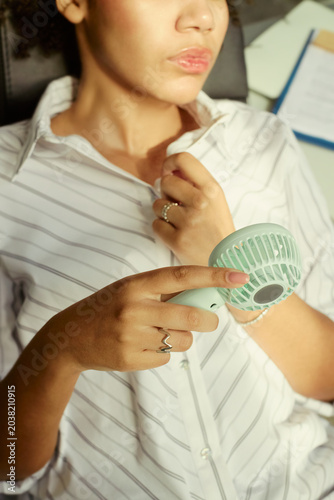 Canvas Print Vertical shot of woman cooling herself with portable fan while loosening collar, showing summer heat discomfort and personal temperature relief