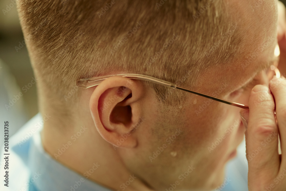 Fototapeta premium Close up of Caucasian young adult man adjusting eyeglasses and rubbing eyes, showing eye strain and fatigue. Suitable for vision care, workplace stress, burnout, or screen fatigue campaigns
