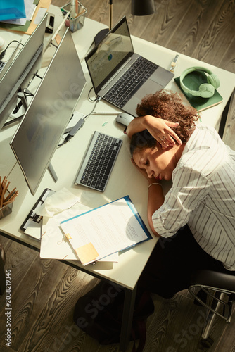 High angle view of young adult African American businesswoman resting head on desk beside laptop and monitor, showing work fatigue and burnout. Useful for stress management or overwork campaigns